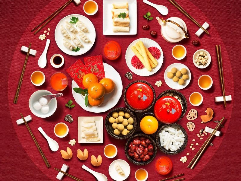 Eine Auswahl an verschiedenen traditionellen Gerichten des chinesischen Neujahrsessen in einem Kreis angeordnet vor einem roten Hintergrund, von oben.