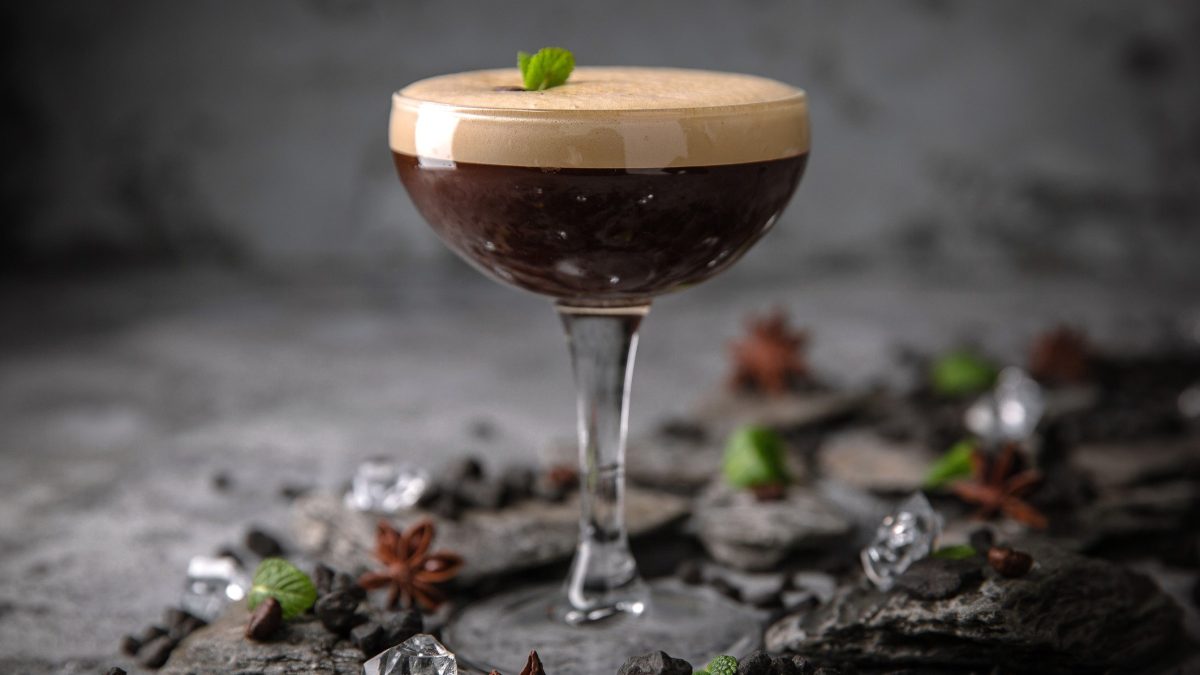 Ein Glas Espresso Martini vor dunkelgrauem Hintergrund, drumherum liegen Eiswürfel und Anissterne.