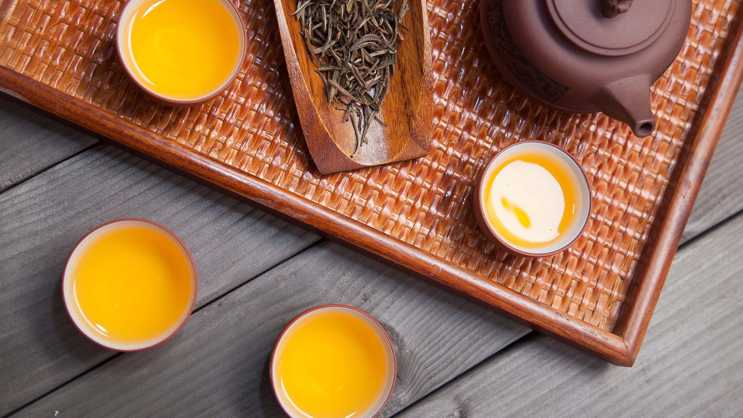 Ein Tablet aus gewobenem Stroh mit einer Ton-Kanne, einem Holzteller mit losen Gelber Tee Blättern und zwei kleinen Tassen mit gelbem Tee, steht neben weiteren zwei Tassen gelben Tee vor einem Holzhintergrund, von oben.