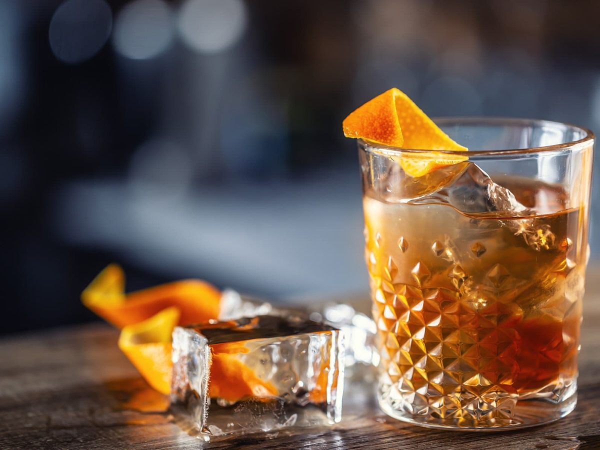 Ein Glas Honey Old Fashioned mit Eis und einer Orangenzeste, daneben liegen ein Eiswürfel und weitere Zesten.
