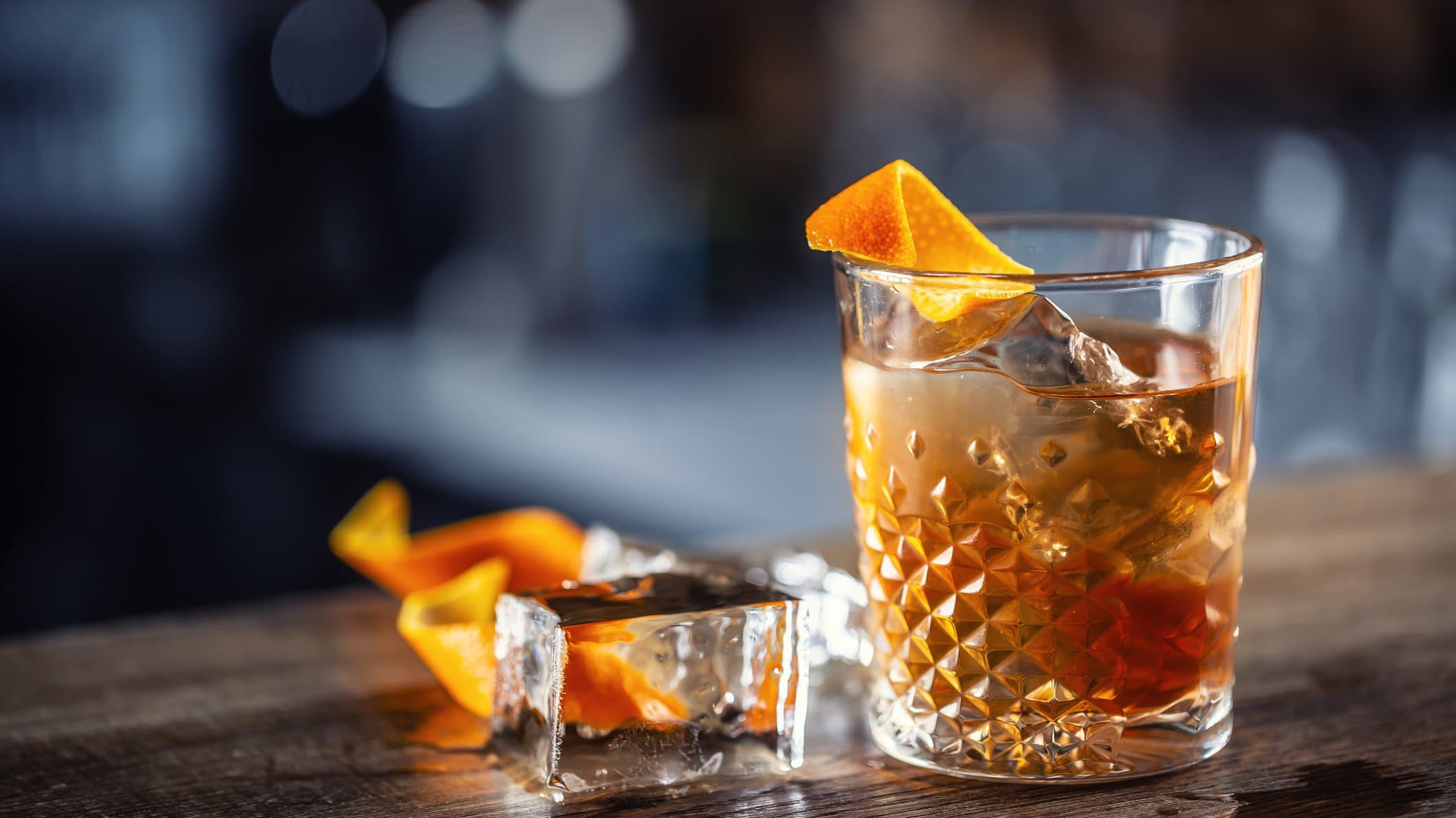Ein Glas Honey Old Fashioned mit Eis und einer Orangenzeste, daneben liegen ein Eiswürfel und weitere Zesten.