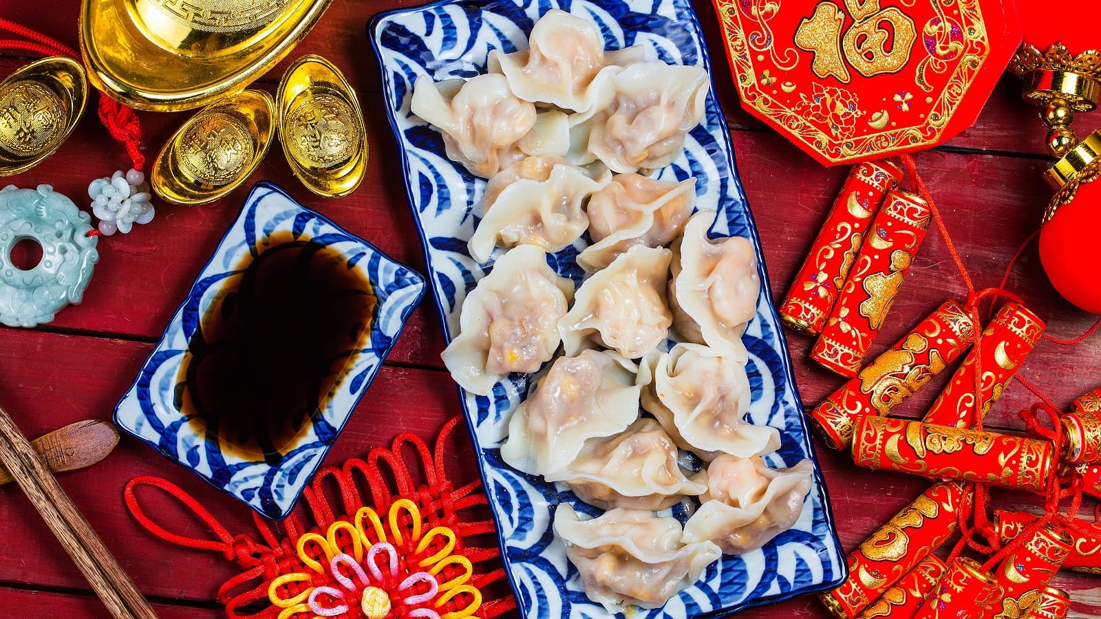 Viele Jiaozi, traditionell chinesische Dumplings auf einem langen Teller angerichtet, neben einer passenden Schale mit Sauce, und vieler roten Neujahrsdekoration vor einem roten Hintergrund, von oben.