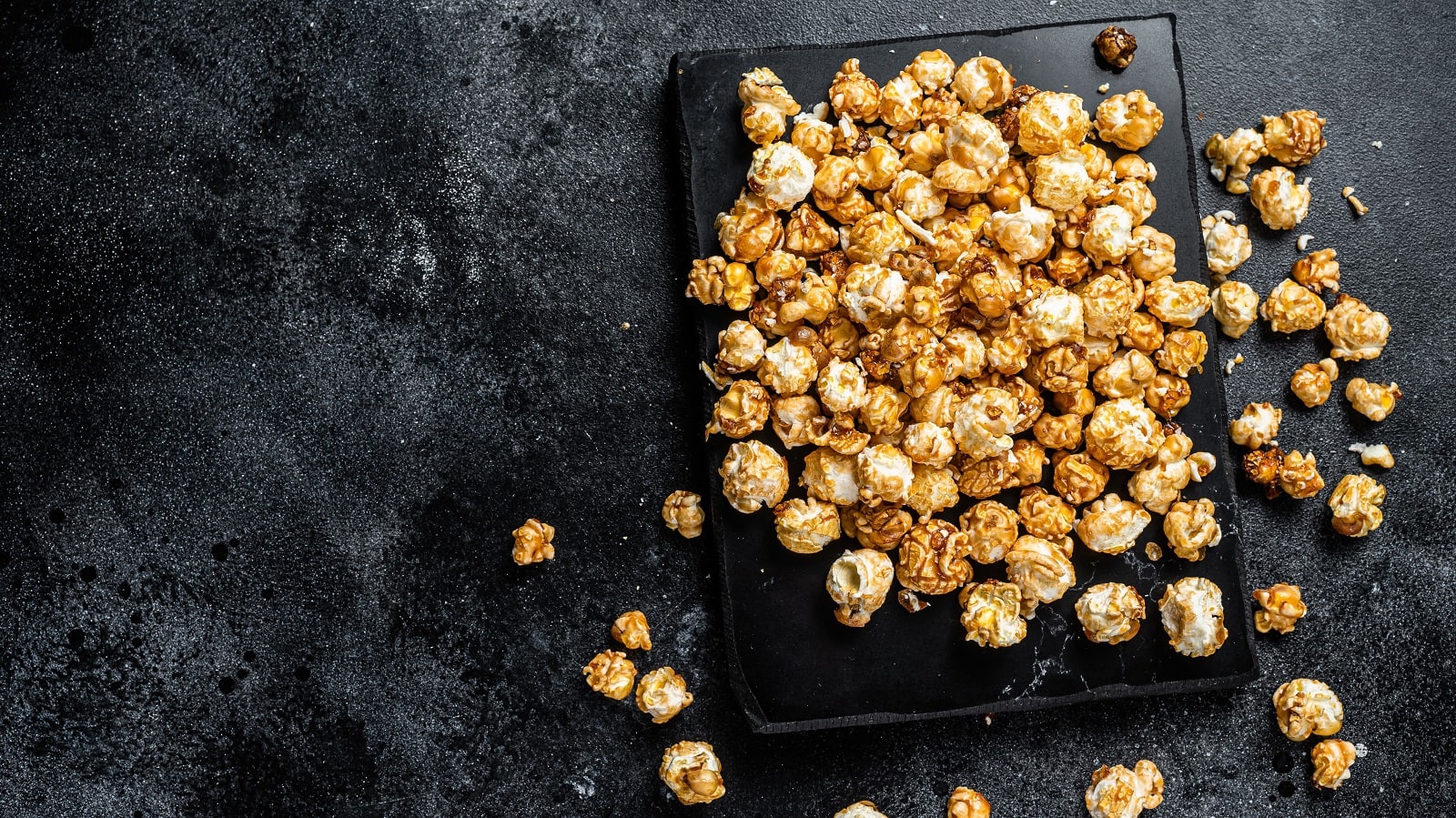 Knuspriges Karamell-Popcorn selbst gemacht im Ofen, auf einem schwarzen Backblech vor einem schwarzen Hintergrund, von oben.