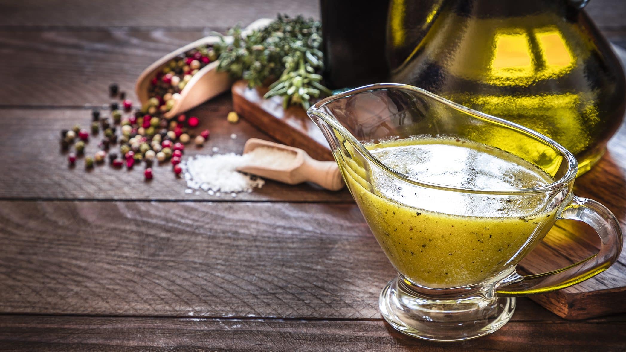 Auf einem Holztisch steht eine Sauciere aus Glas. In dieser ist eine klassische Vinaigrette. Dabei handelt e sich um ein Salatdressing. Zu sehen sind auch eine Glasflasche mit Olivenöl, Pfefferkörner und Kräuter. Sowie ein Holzlöffel mit Salz.
