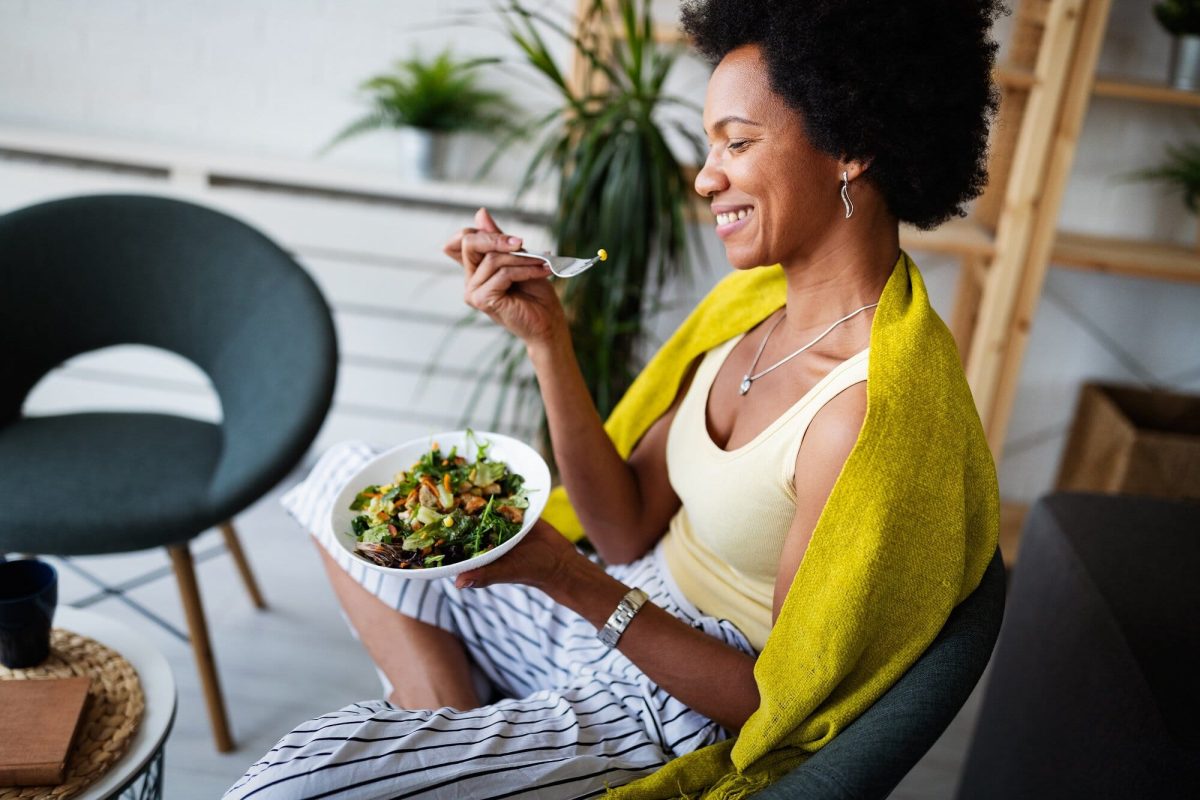 Langsam und bewusst essen. Eine Frau in einer gestreiften Hose und einem gelben Top sitzt gemütlich auf einem Stuhl und isst einen frischen Salat.