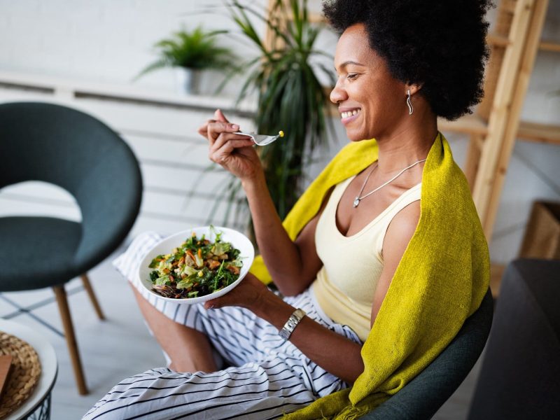 Langsam und bewusst essen. Eine Frau in einer gestreiften Hose und einem gelben Top sitzt gemütlich auf einem Stuhl und isst einen frischen Salat.