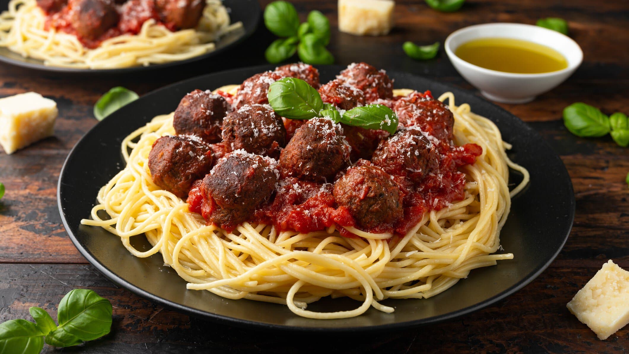 Ein Teller Spaghetti mit veganen Fleischbällchen, daneben liegen Basilikumblätter und ein paar Stücke Parmesan.