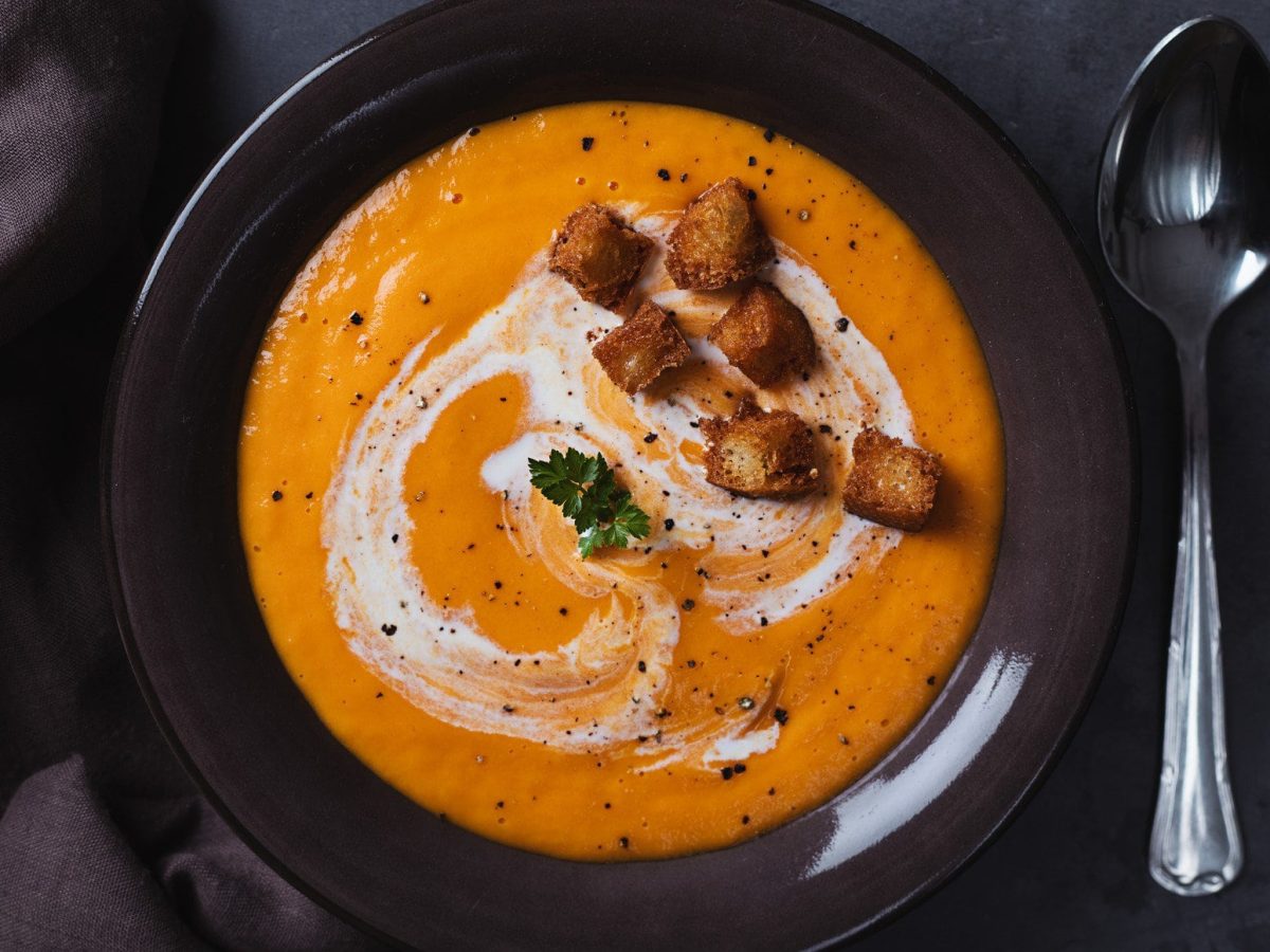 Eine orangefarbene Suppe mit Zimt-Croutons in der Draufsicht, daneben liegt ein Löffel.