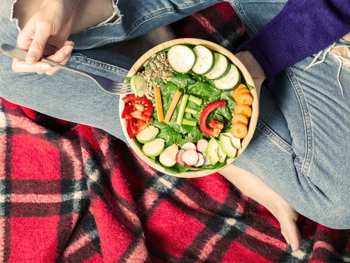 Eine Frau sitzt oim Schneidersitz auf einer Decke und hält eine Gemüse-Bowl in der Hand, in der unter anderem das Wort "Veg" mit Gemüse gelegt wurde.