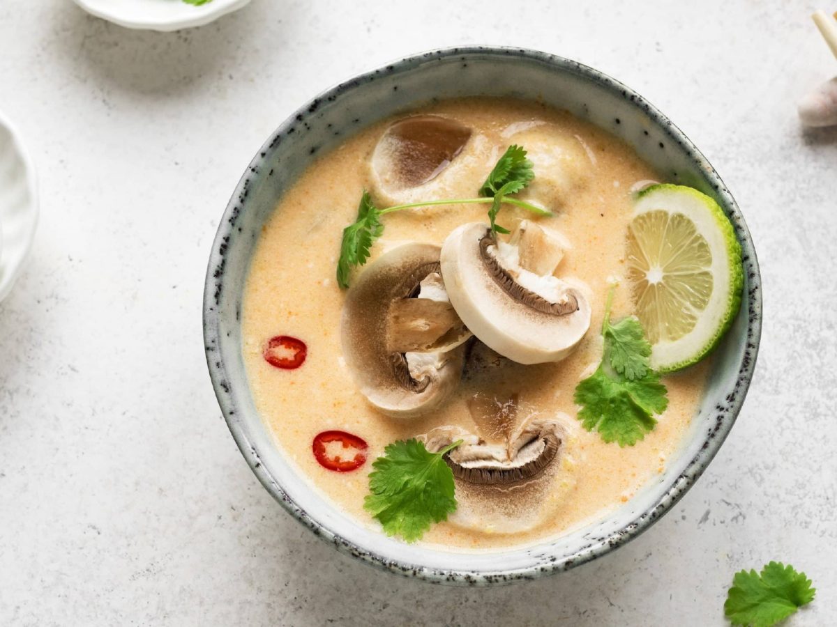 Tom Kha Hed, vegane thailändische Suppe mit Kokosmilch, Zitronengras, Ingwer, Pilzen, Chili, Limette und Chili in einer hellen Schale auf weißem Hintergrund