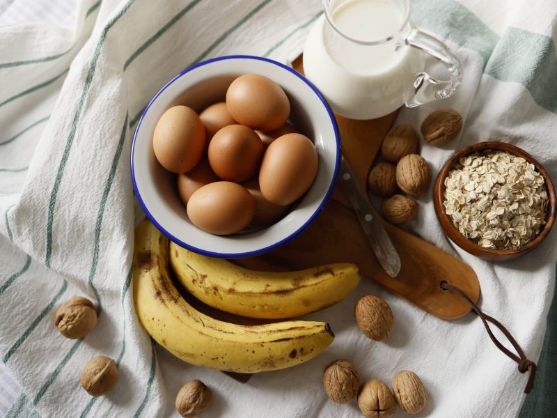 Auf Geschirrtüchern stehen eine Schale mit Eiern, eine mit Haferflocken, eine Kanne Milch, außerdem sieht man Nüsse und Bananen.