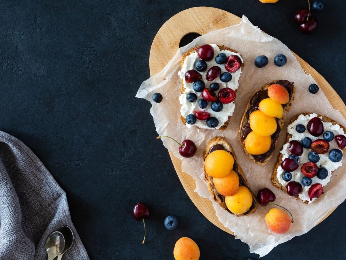 Drei Scheiben mit vitaminreichem Frühstücksbrot liegen auf einem ovalen Tablett auf einem dunklen Untergrund. Die Brote sind angerichtet mit Hüttenkäse, Schokocreme, Aprikosen, Kirschen und Heidelbeeren. Ein wirklich gesundes Frühstück.