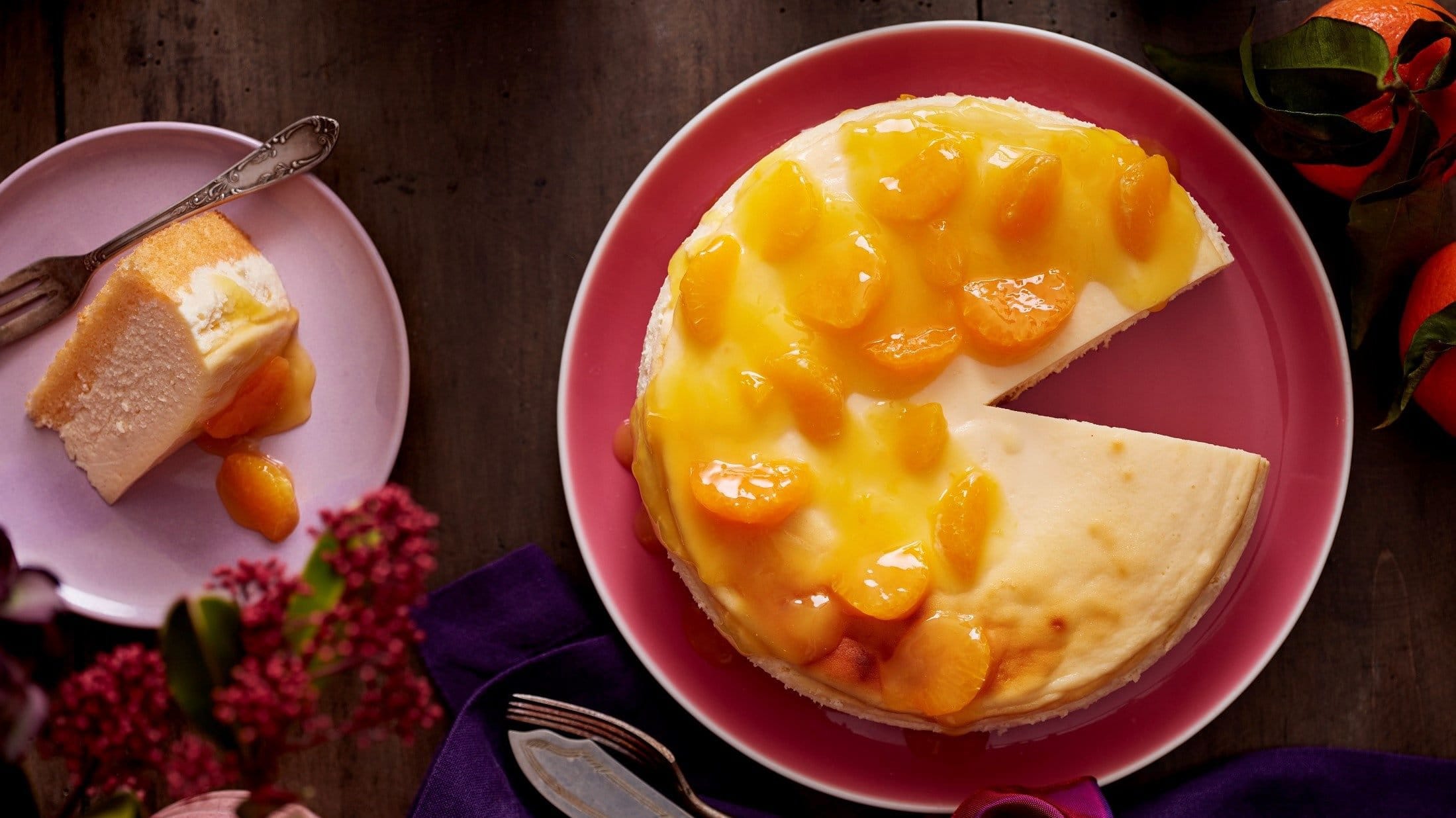 Unser Mandarinen-Cheesecake liegt angeschnitten auf einem Teller. Ein einzelnes Stück liegt auf einem zweiten Teller links daneben.