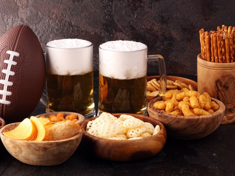 Zwei Bier, verschiedene Snacks und ein Football vor dunklem Hintergrund