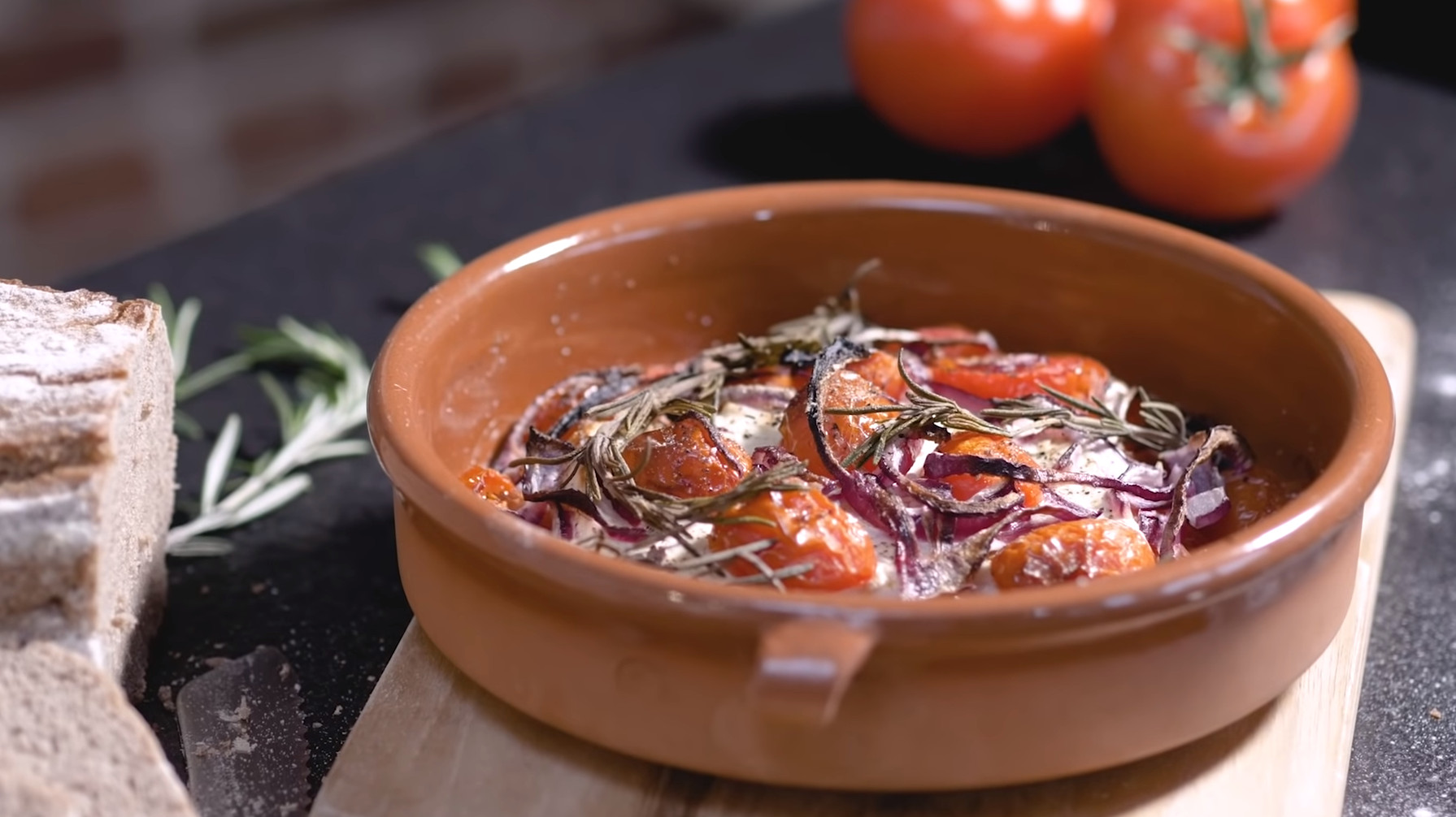 gebackener Feta mit Tomaten in Terracotta-Schale auf einem Holzbrett.