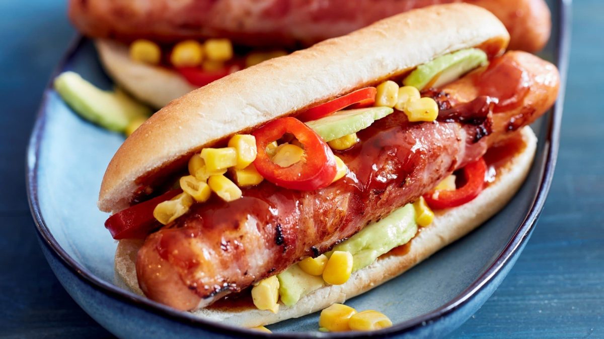 Hot Dogs mit Mais und Avocado auf einem blauen Teller. Das Ganze auf einem blauen Untergrund.