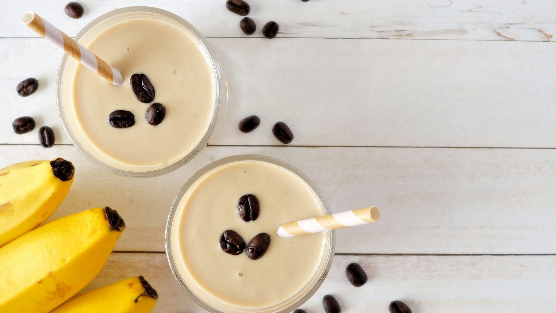 Zwei Gläser Kaffee-Bananen-Smoothie stehen auf einem weißen Holztisch. Kaffeebohnen und Bananen als Deko dazu.