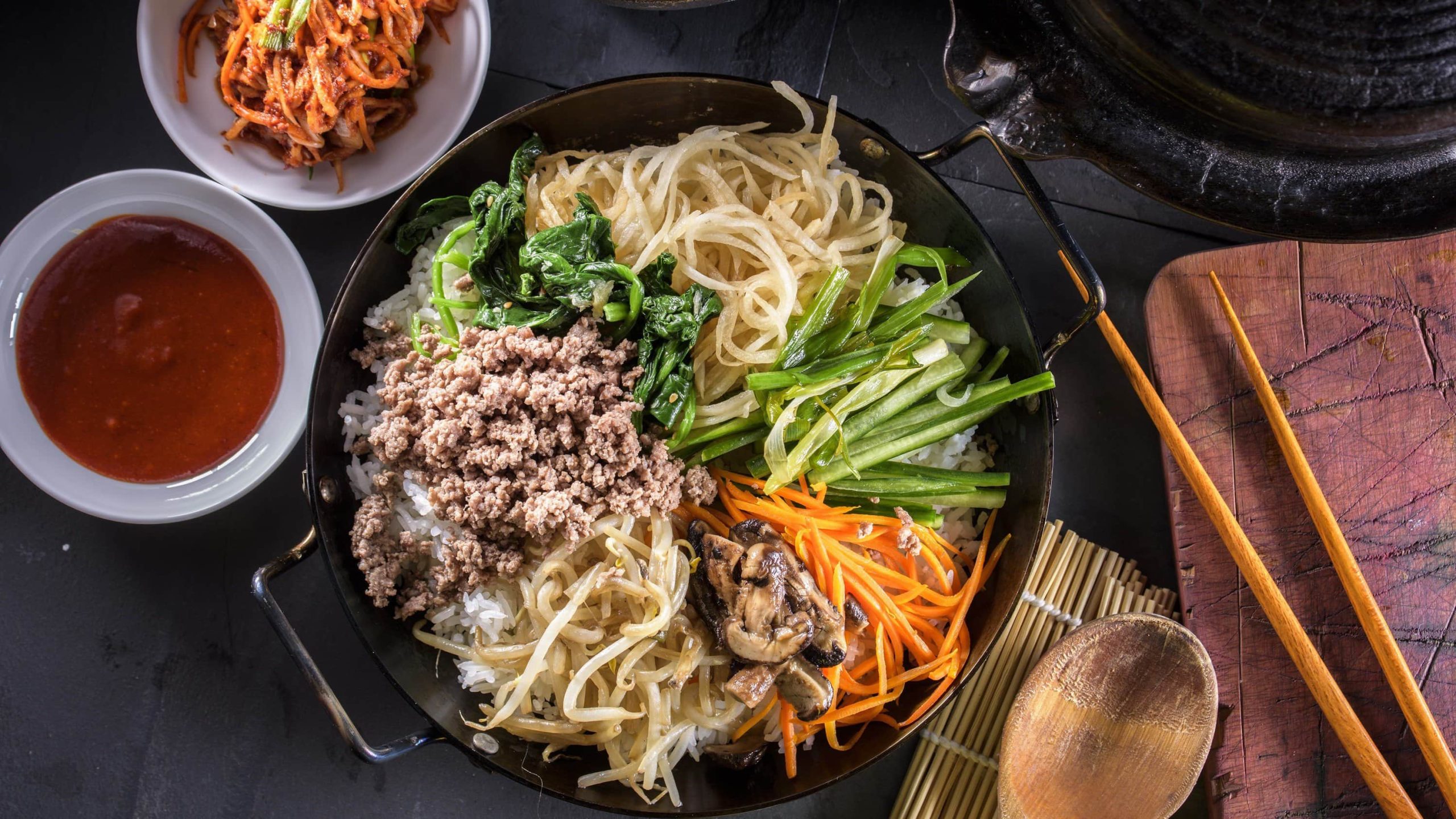 Koreanisches Bibimbap, bestehend aus Karotten, Pilzen, Rettich, veganem Hackfleisch, Gurke, Karotte und Sprossen auf einem Bett aus Reis in einer schwarzen Schale, dazu Chili-Sauce und Kimchi