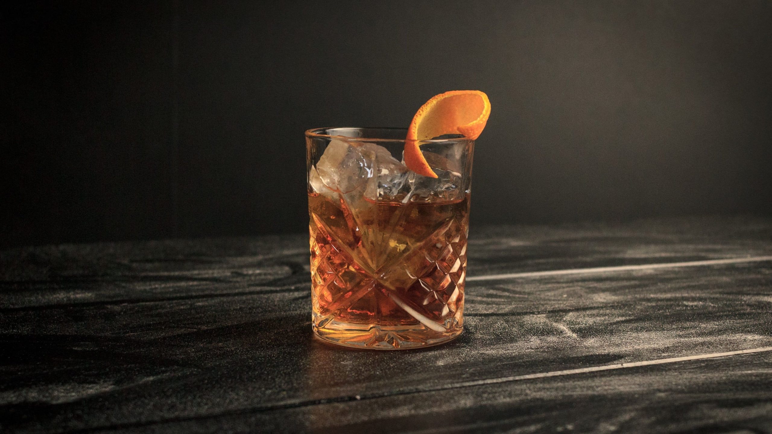 Ein Glas Old Fashioned mit einer Orangenzeste und Eis vor dunklem Hintergrund.