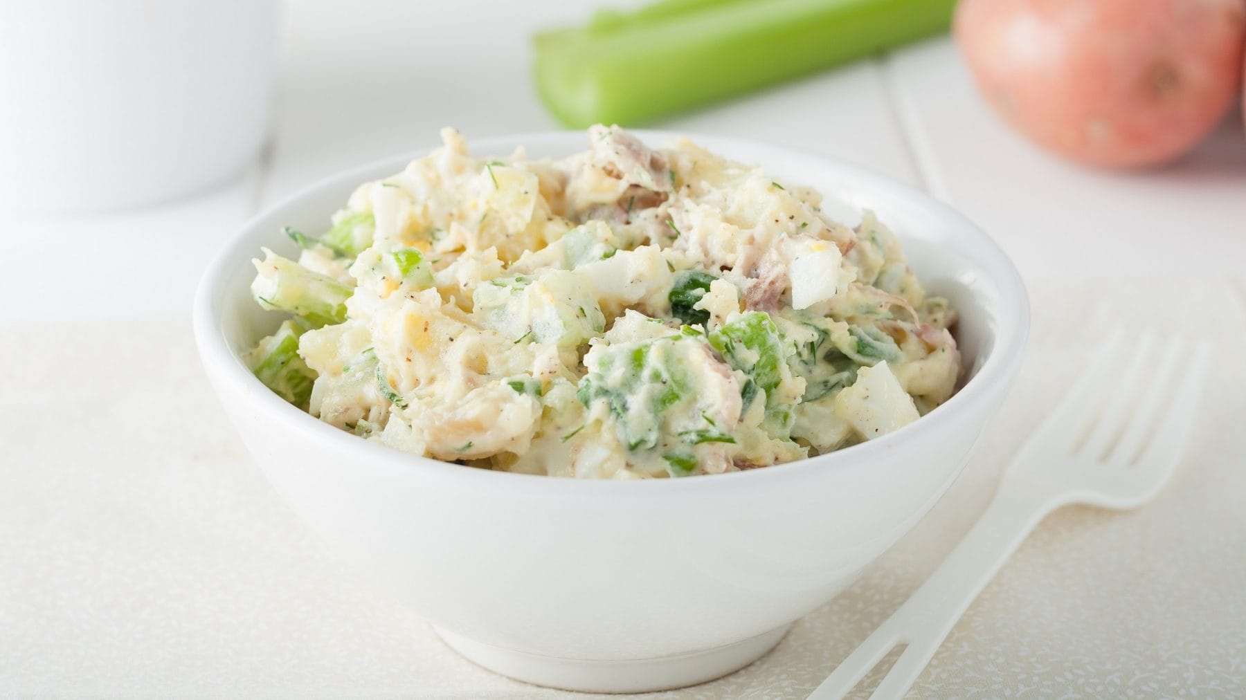 In einer weißen Schale ist ein Sellerie-Kartoffel-Salat. Daneben liegen eine weiße Gabel, eine Kartoffel und eine Sellerie-Stange.