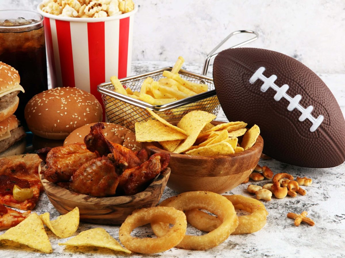 Super Bowl 2024 Tisch gedeckt Pommes, Nachos, Pizza, Burger, Chicken Wings, Popcorn, einem Glas Kola und einem Football. Angerichtet auf einem hellen Steintisch. Frontalaufnahme.