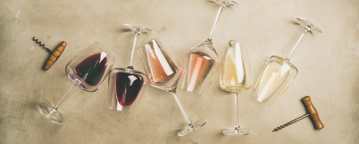 Sechs verschieden gefüllte Weingläser liegen auf einer grauen Betonwand. Links und rechts daneben liegt jeweils ein Korkenzieher.