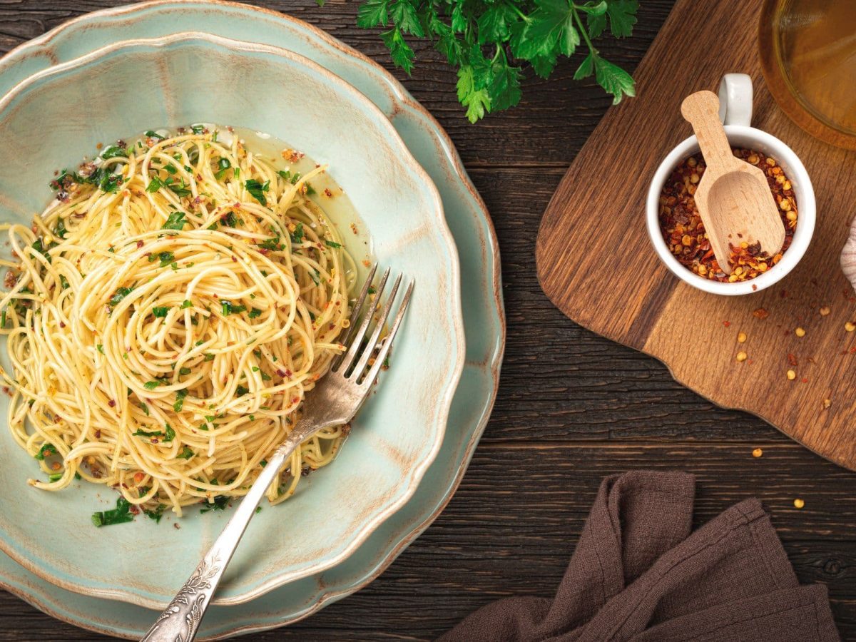 Spaghetti Aglio, Olio e Peperoncini auf rustikalem Teller. Das Ganze steht auf einem dunklen Holz und ist umgeben von den Hauptzutaten Chili, Knoblauch und Co