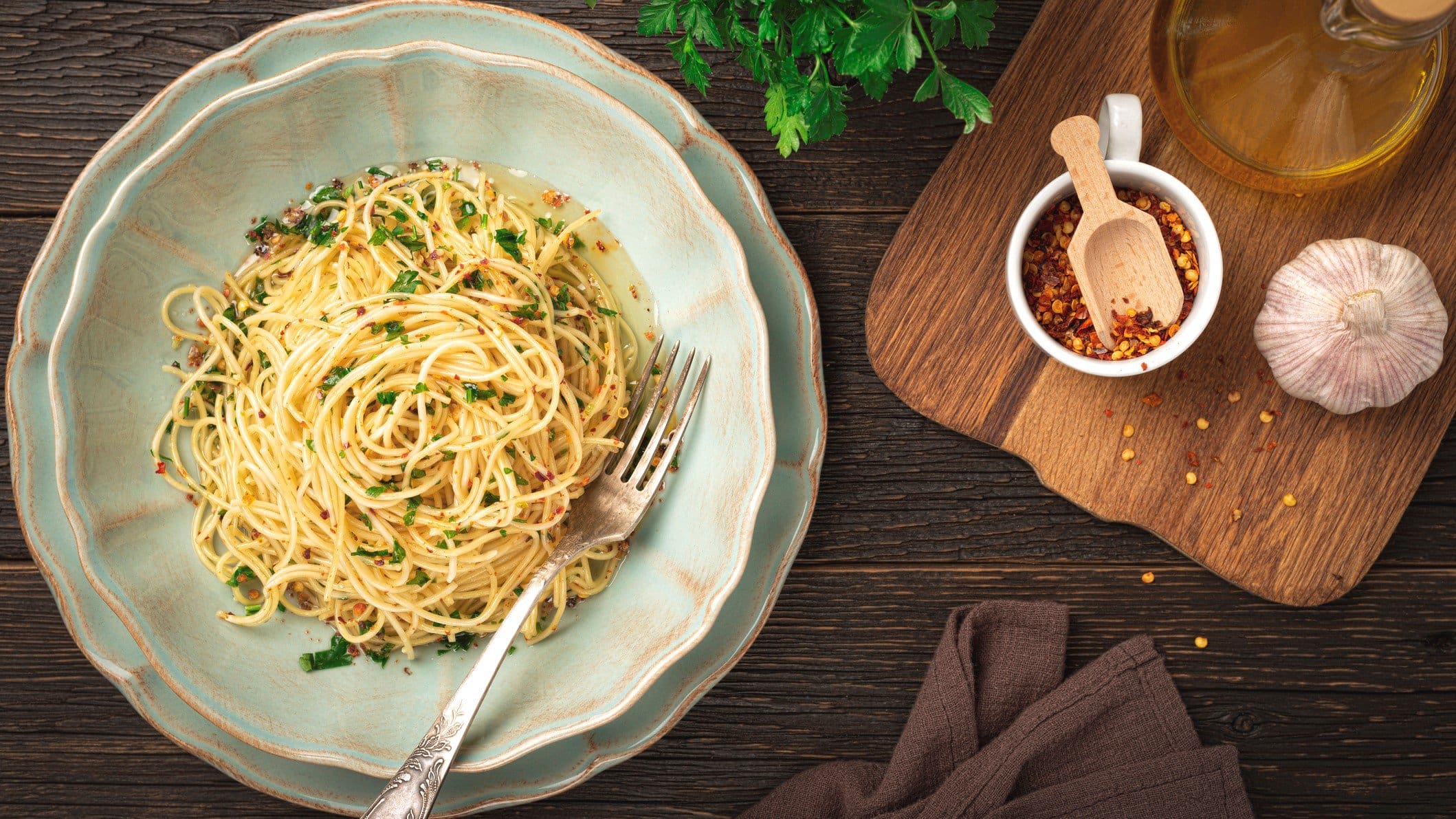 Spaghetti Aglio, Olio e Peperoncini auf rustikalem Teller. Das Ganze steht auf einem dunklen Holz und ist umgeben von den Hauptzutaten Chili, Knoblauch und Co