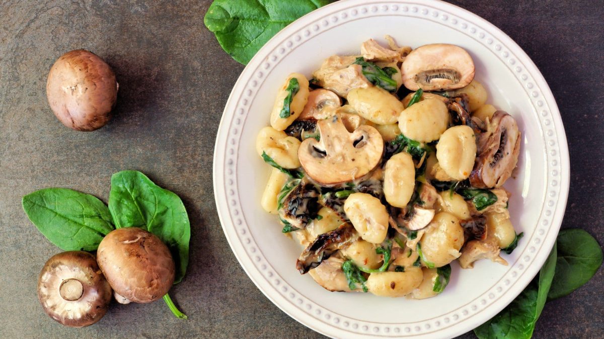 Gnocchi-Pfanne mit Pilzen und Spinat auf weißem Teller, angerichtet auf grauem Untergrund. Umgeben ist der Teller von Champignons und Spinat.