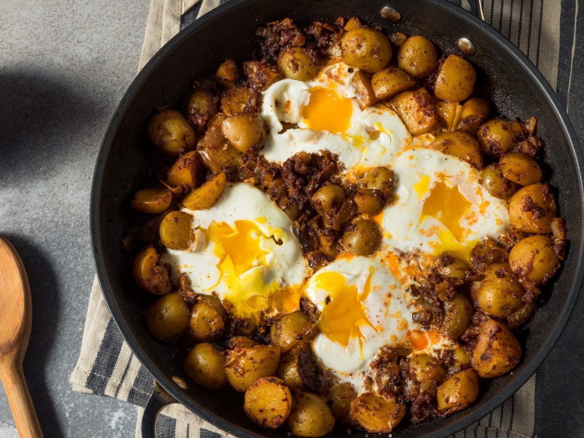 Huevos Rotos, spanische zerbrochene Eier, mit Kartoffeln und Chorizo in einer Pfanne. Diese steht auf grauem Untergrund.
