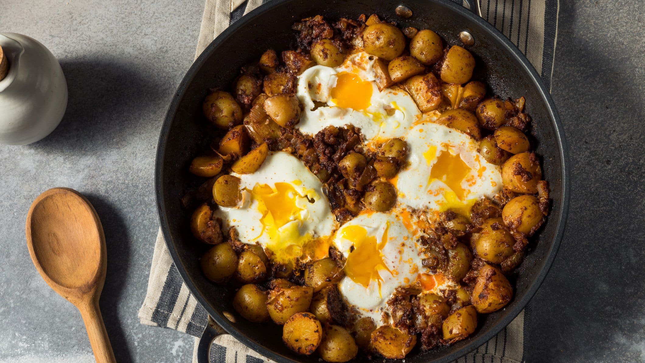 Huevos Rotos, spanische zerbrochene Eier, mit Kartoffeln und Chorizo in einer Pfanne. Diese steht auf grauem Untergrund.