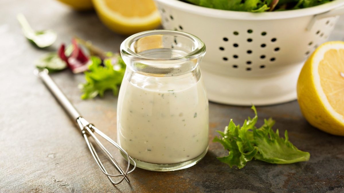 Joghurt-Dressing vor Sieb mit Salat auf grauer Oberfläche. Daneben liegen ein Schneebesen, einige Salatblätter und angeschnittene Zitronen.