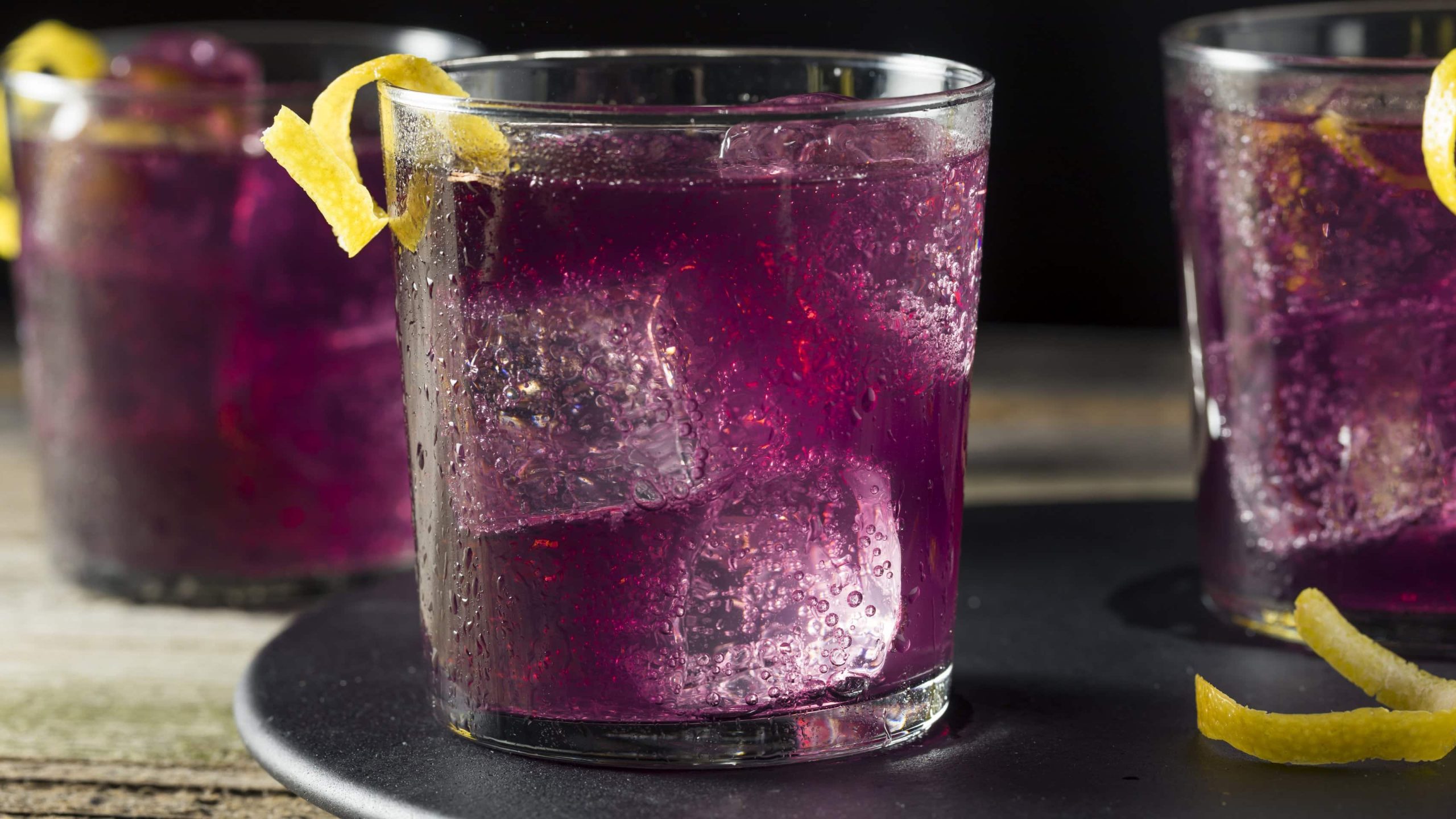 Violetter Cocktail auf schwarzem Teller mit einem Zitronenstreifen garniert.