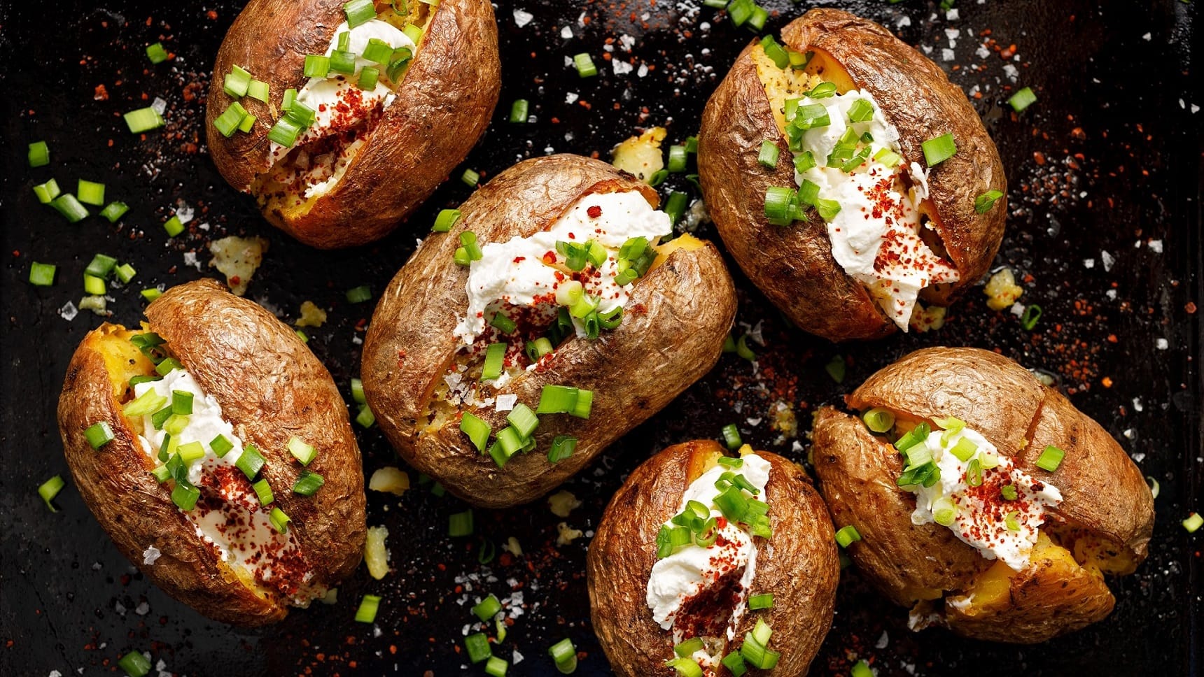 Backkartoffeln mit Bärlauchquark, Frühlingszwiebeln und frisch gemahlener Pfeffer auf dunklem Untergrund in der Draufsicht.