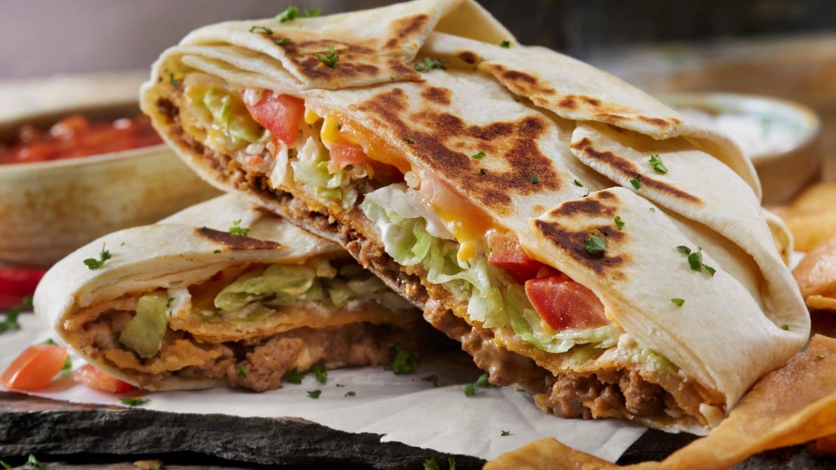 Halbierter Crunch Wrap Surpreme von Taco Bell übereinander auf Krepp auf dunklem Untergrung. Daneben Nachos und im Hintergrund weißer und roter Dip. Frontalansicht.