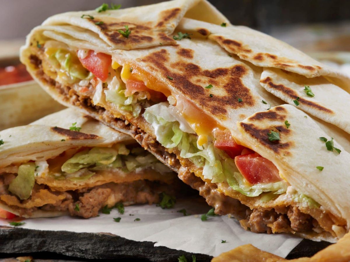 Halbierter Crunch Wrap Surpreme von Taco Bell übereinander auf Krepp auf dunklem Untergrung. Daneben Nachos und im Hintergrund weißer und roter Dip. Frontalansicht.