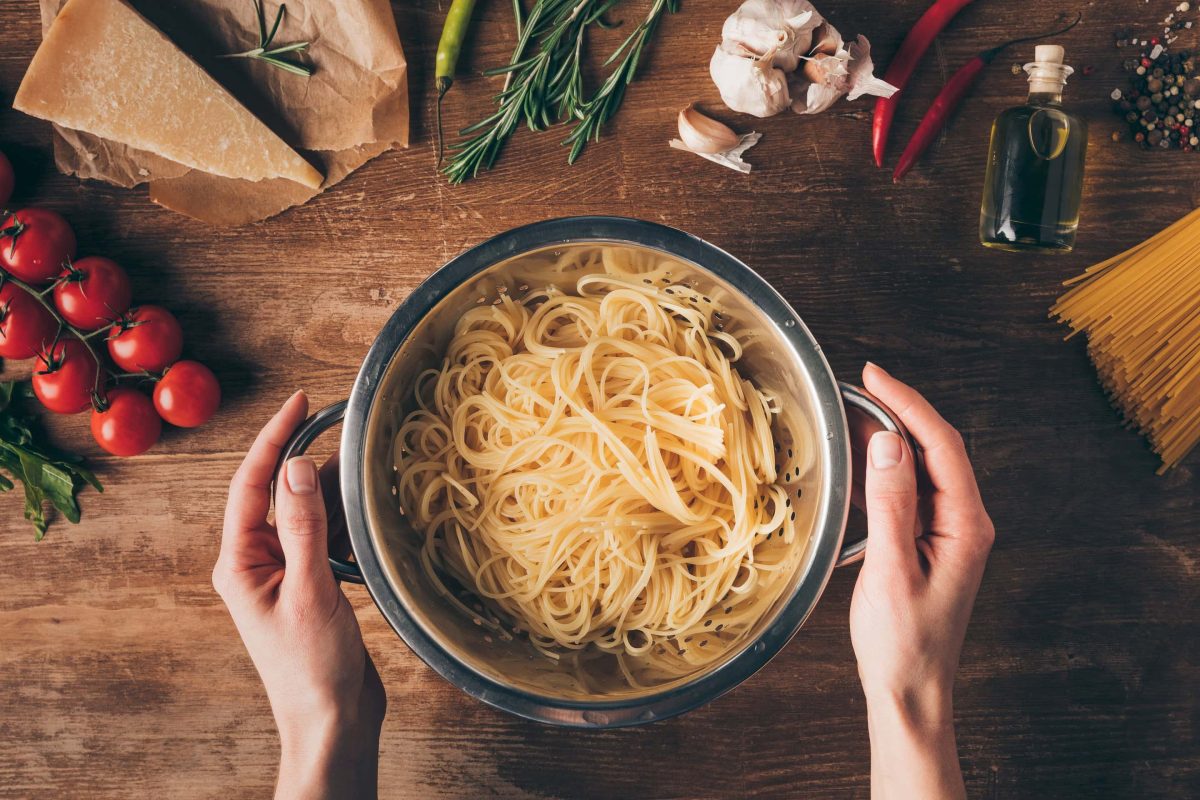 Ein Topf Spaghetti in den Händen einer Frau in der Draufsicht, drumherum liegen auf einem Holztisch Gemüse, Parmesan, Nudeln und andere Zutaten.