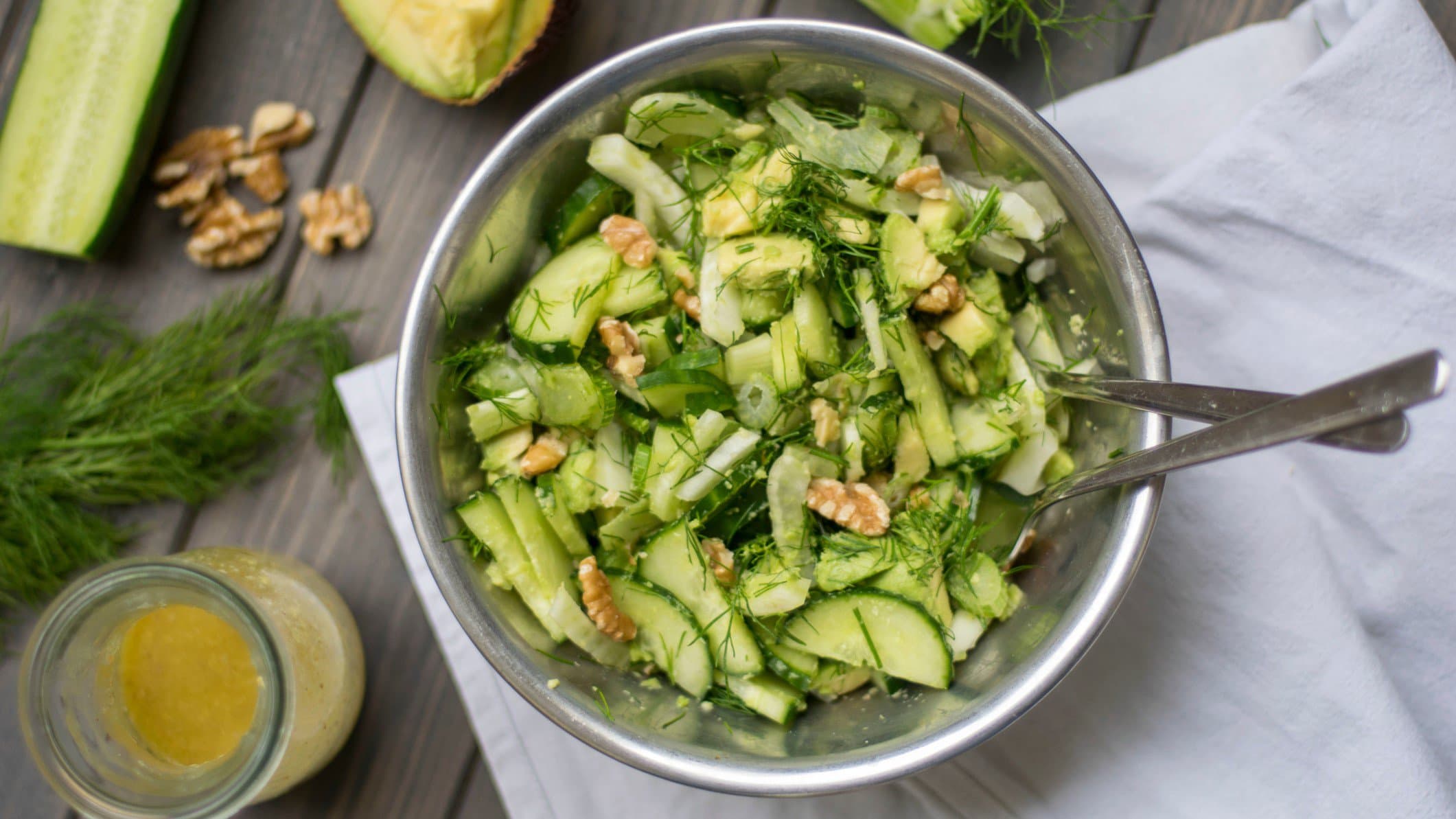 Eine Schüssel Gurkensalat mit Fenchel und Walnüssen mit Salatbesteck in der Draufsicht, daneben Zutaten wie Nüsse, Fenchelgrün, Gurke und Dressing.