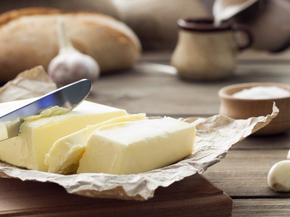 Auf einem Holztisch liegen Butter im Packpapier, zwei Knoblacuzehen, Eier, Brot und eine Schale mit einer weißen Masse drin.
