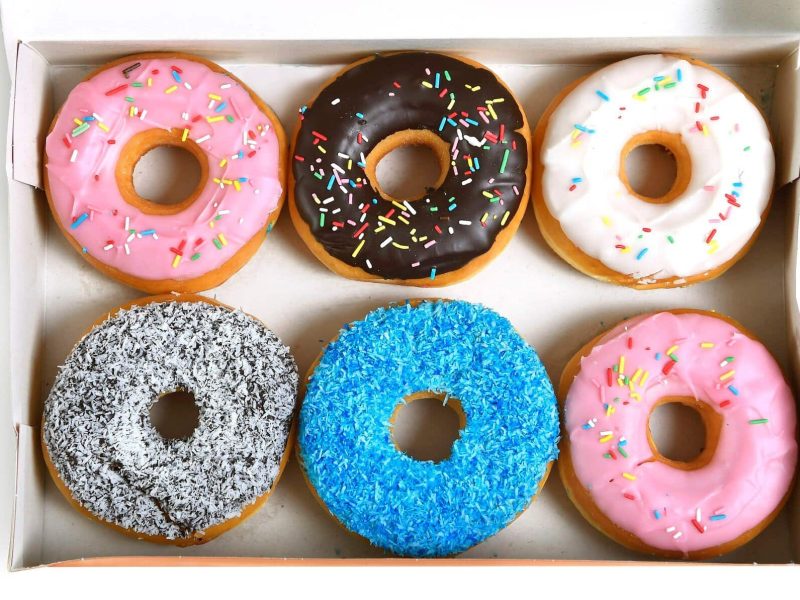 Sechs verschiedene Donuts liegen in einer weißen Box auf einem weißen Untergrund. Zwei Donuts sind mit rosa Glasur überzogen, einer mit blauen Streuseln, einer mit Schokolade, ein weiterer Donut mit Schokolade und Kokosraspeln und ein letzter mit weißem Zuckerguss.
