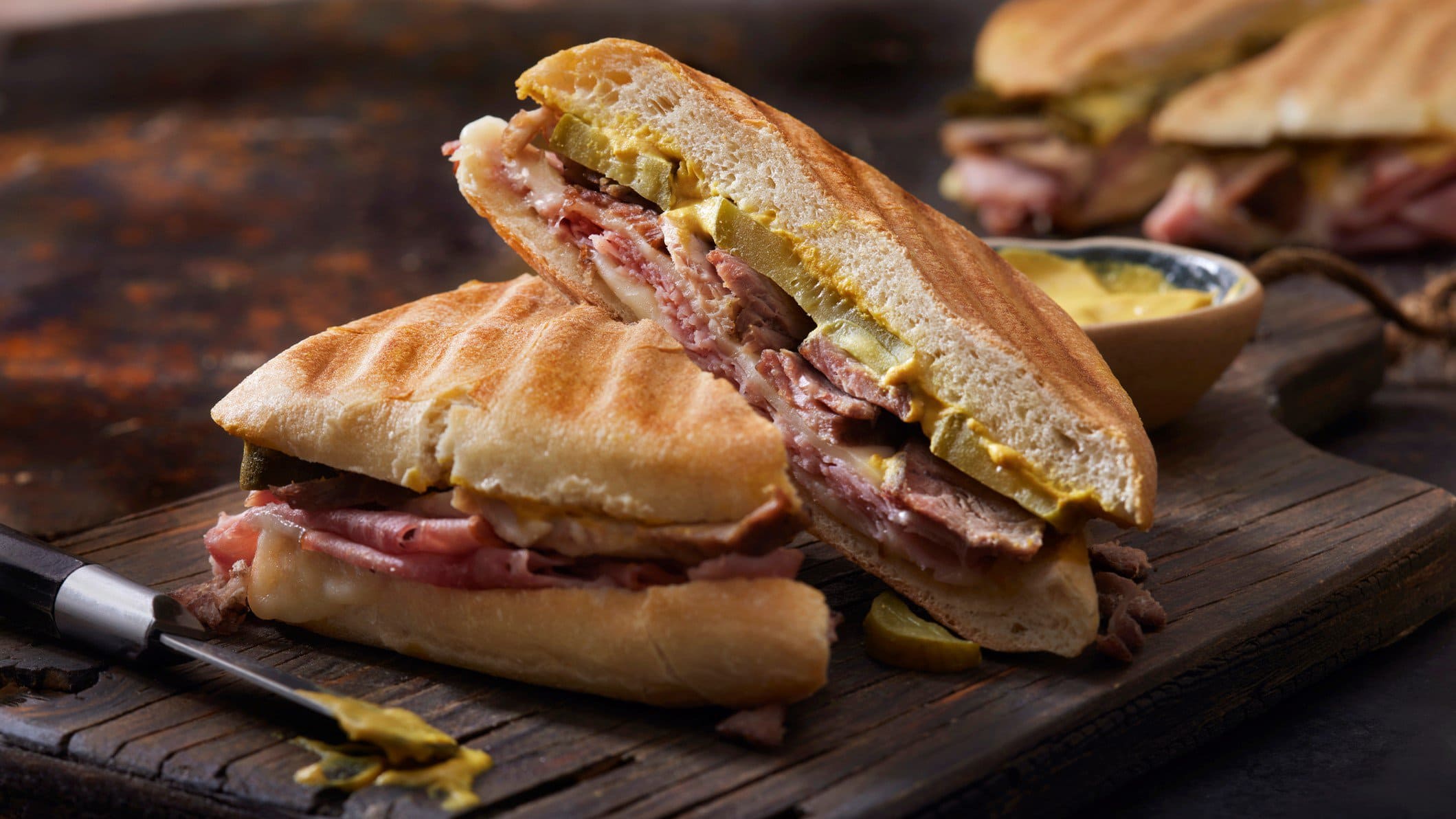 Zwei grilled Cuban Sandwiches auf dunklem Holzbrett auf Holztisch. Messer mit schwarzem Griff und Senf im Vordergrund. Rechts unscharf weiteres Sandwich. Frontalansicht.