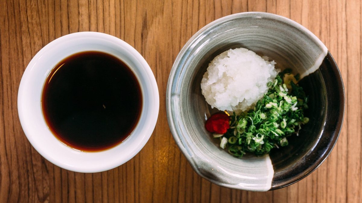 Ponzu-Sauce in weißer Schale neben Schale mit Reis, Hackfleisch und Gemüse auf Holztisch. Aufnahme aus der Vogelperspektive.