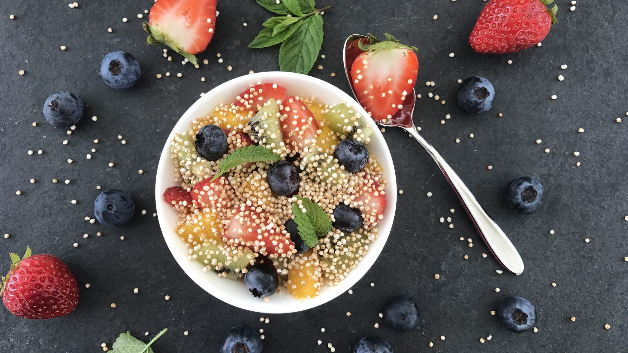 Unser Quinoa-Obstsalat steht angerichtet in einer Schüssel auf einem schwarzen Hintergrund, umringt von Erdbeeren und Blaubeeren.