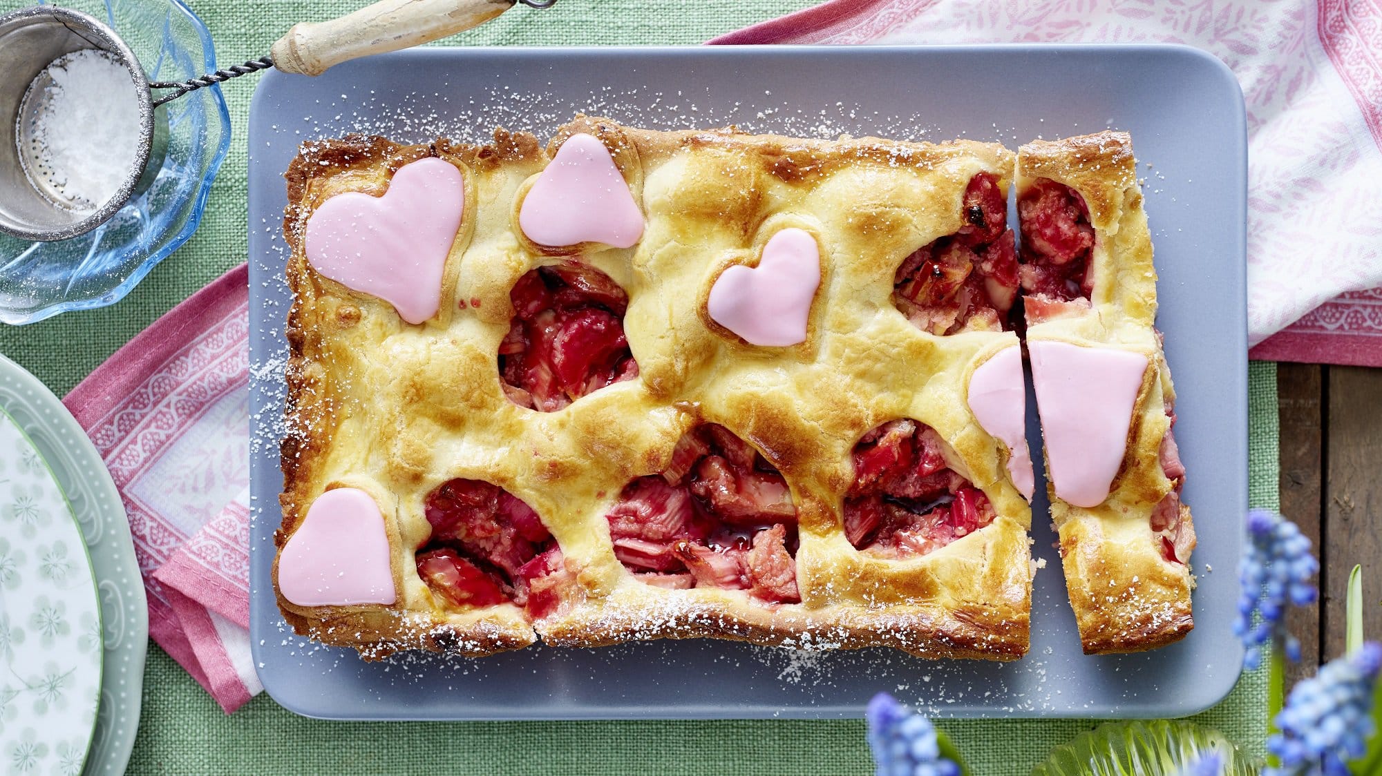 Ein Rhabarber Pie mit Herzen steht mit Puderzucker bestreut auf einer hellen Form. Aus der Teigdecke sind einige Herzen ausgeschnitten. Weitere rosa Herzen sind auf dem Pie verteilt.
