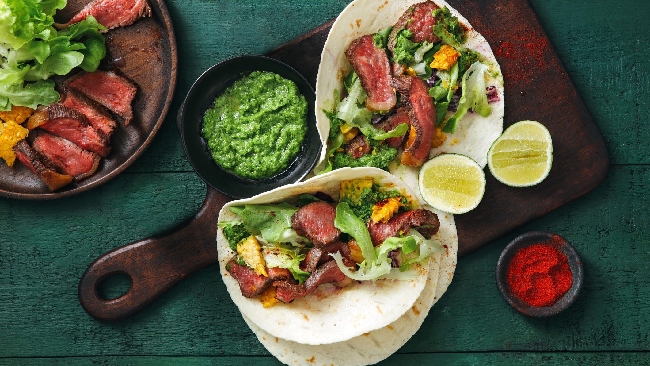 Steak Tacos mit grünem Pesto auf einem Holzbrett. Daneben verschiedene Zutaten auf einem blauen hölzernen Untergrund.