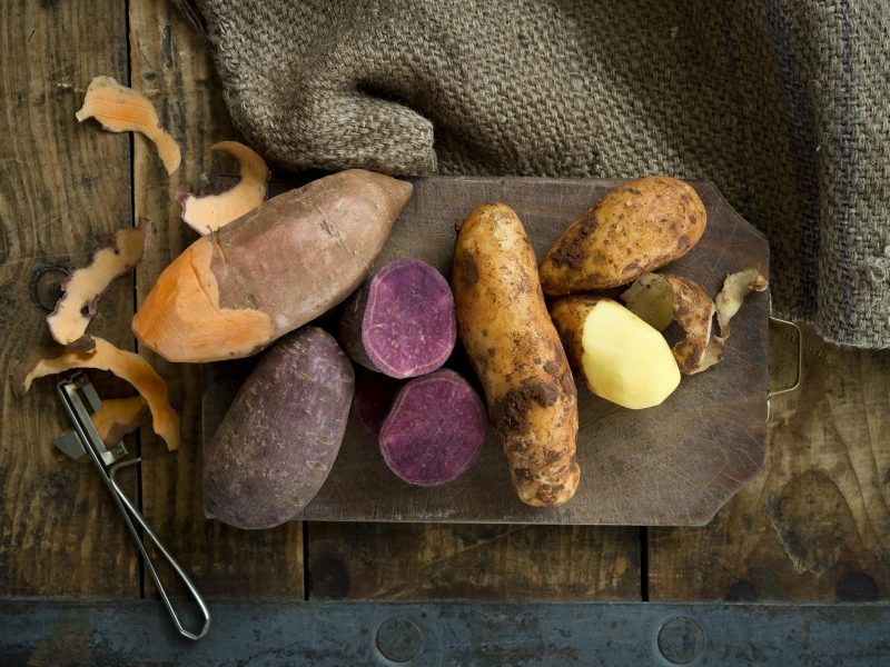 Verschiedene Kartoffel- und Süßkartoffelsorten auf einem Brett auf Holzboden in der Draufsicht, daneben ein Sparschäler.