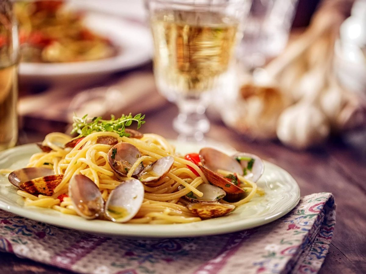 Seitliche Aufnahme einer Portion Spaghetti alle Vongole auf einem Holztisch mit Weinglas und Thymian im Hintergrund.