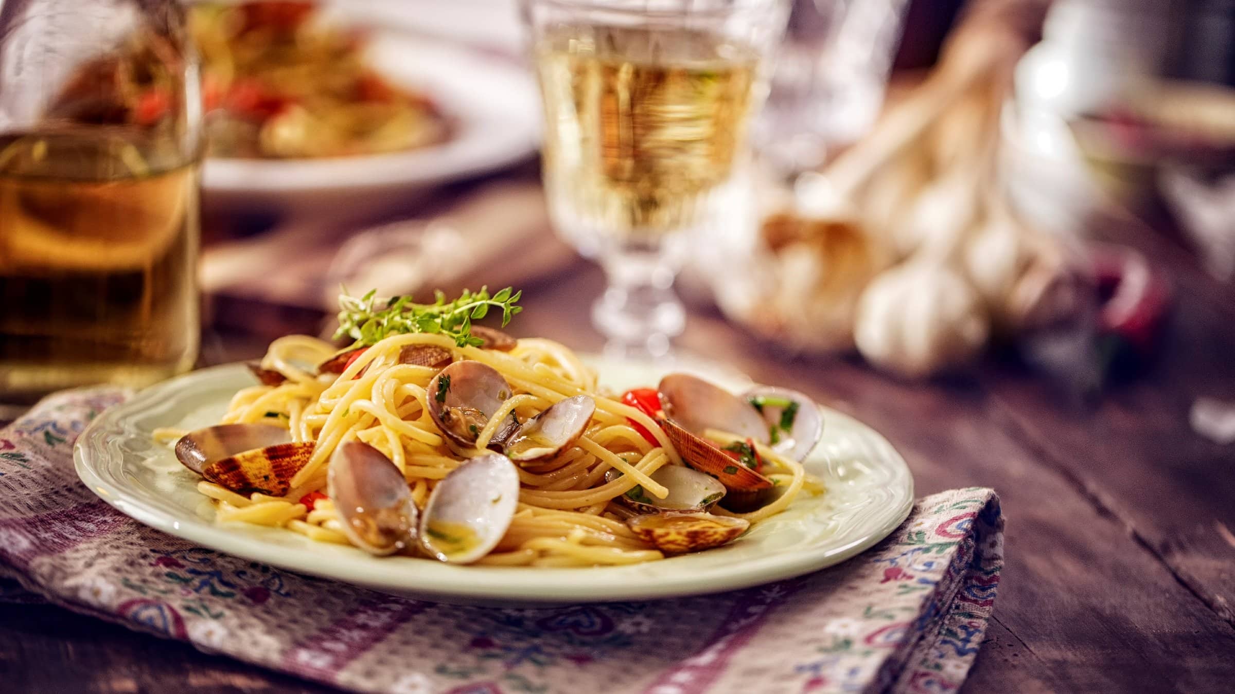 Seitliche Aufnahme einer Portion Spaghetti alle Vongole auf einem Holztisch mit Weinglas und Thymian im Hintergrund.