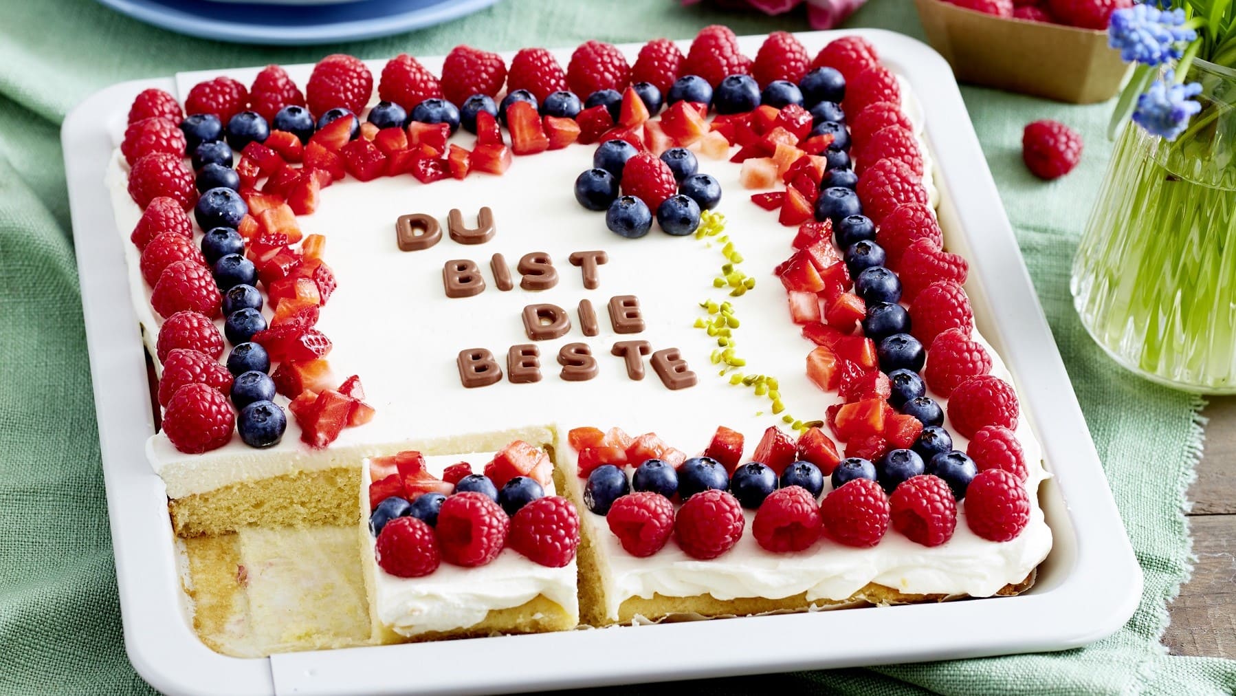 Auf einem Blech liegt ein Du-bist-die-Beste-Kuchen. Er ist dekoriert mit frischen Beeren und essbaren Buchstaben.