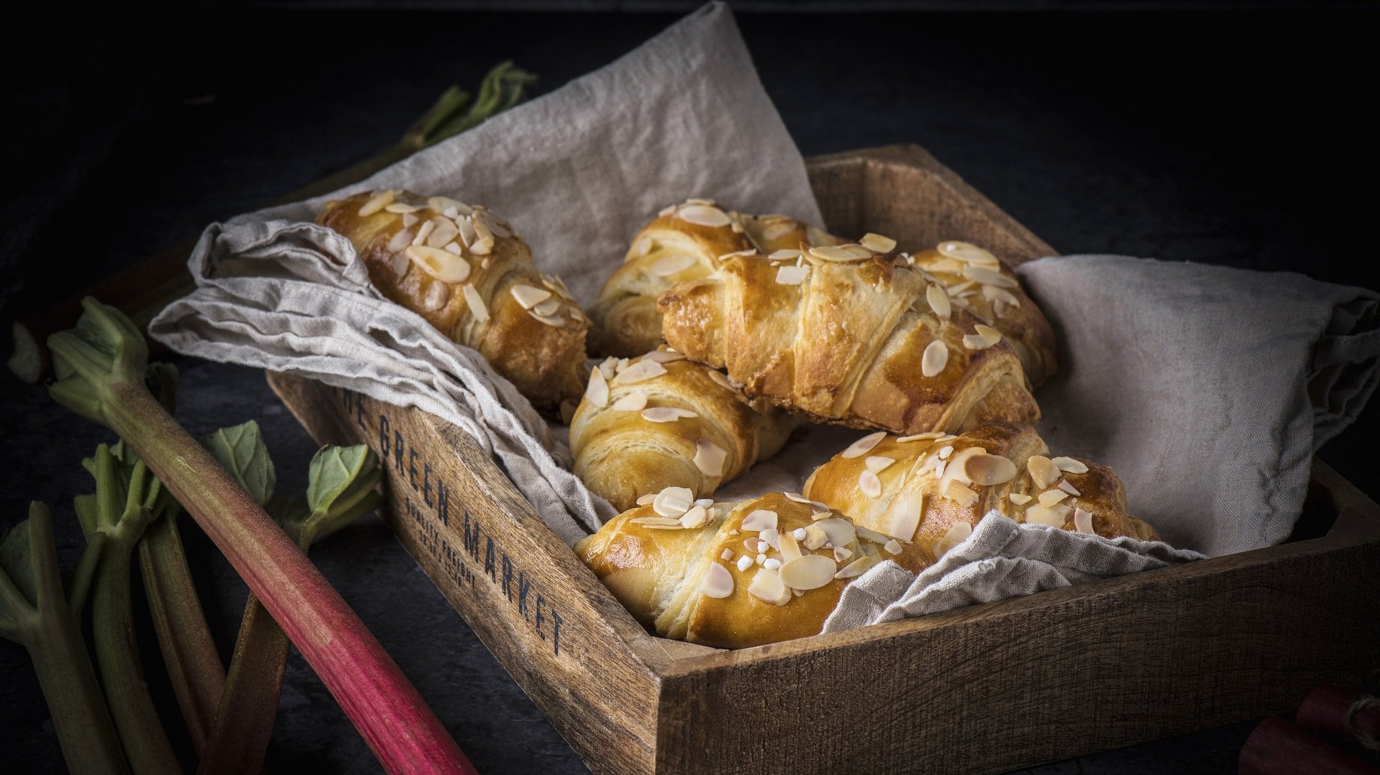 In einem Brotkorb liegen mehrere Rhabarber-Croissants. Sie sind mit Mandelblättchen bestreut und sind goldbraun gebacken.